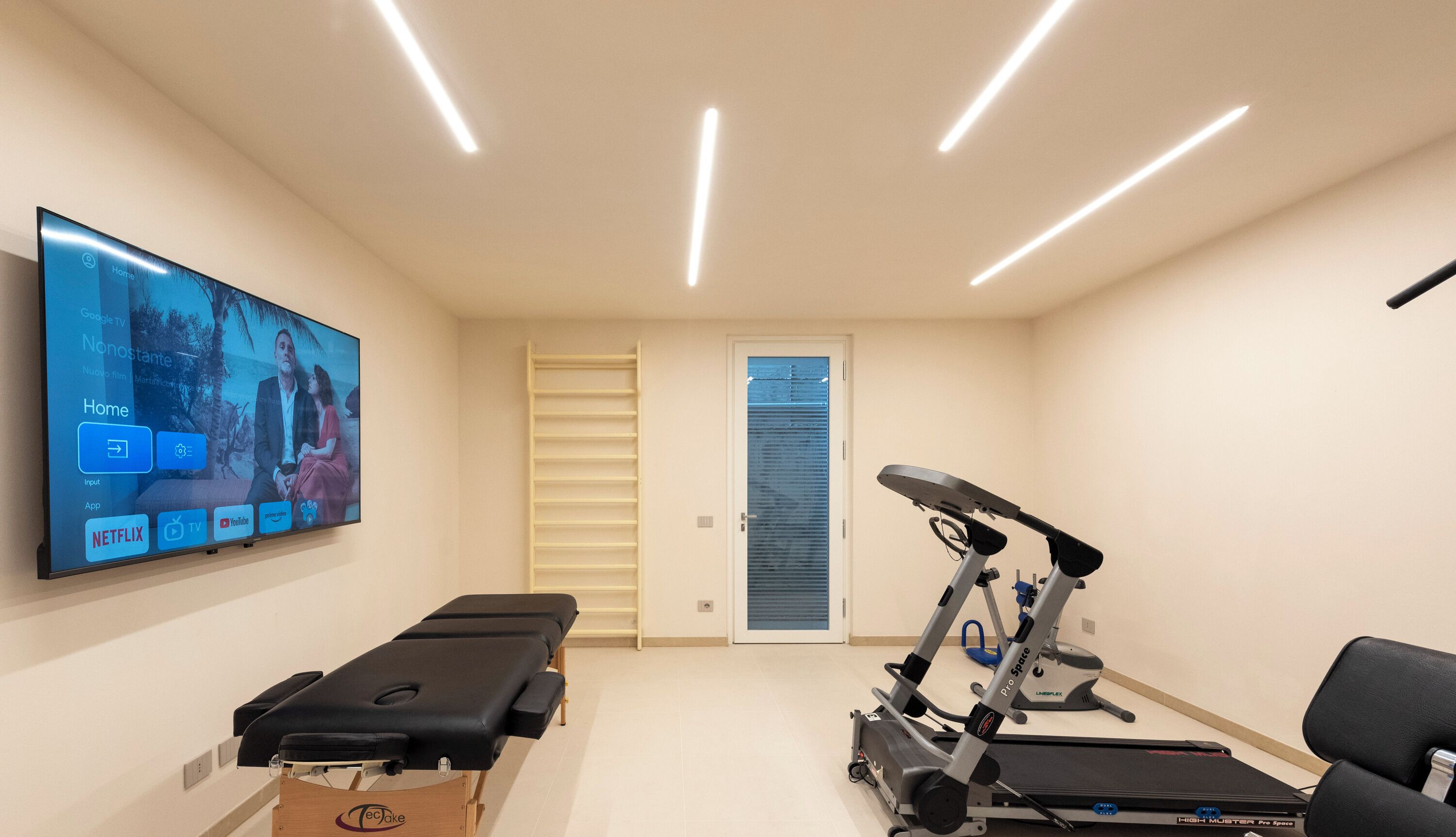 Une pièce équipée d’un tapis de course, d’une table de massage et d’un téléviseur affichant un écran d’accueil.