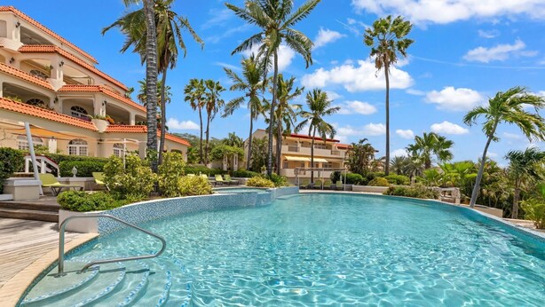 A clear blue pool with a tiled edge, surrounded by palm trees and a building with a red-tiled roof. - Palm Piscadera · Ocean Views · Pool · Beaches (Piscadera)