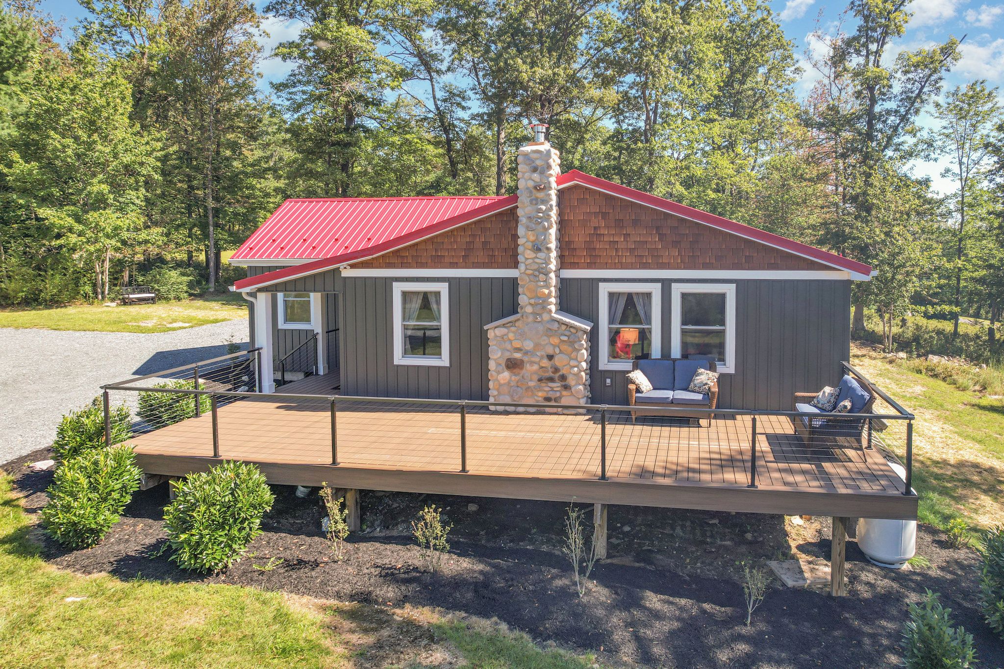 Une cabane avec un toit rouge, une cheminée en pierre et une terrasse dotée de meubles d’extérieur.