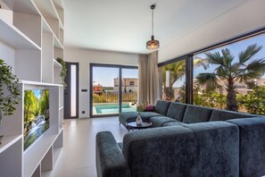 Una sala de estar moderna con un sofá grande, una mesita y vistas a una piscina y palmeras.