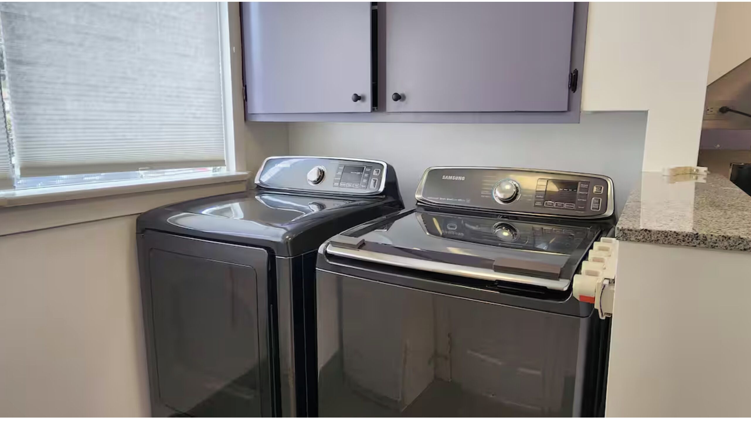 A laundry area with two front-loading washing machines and a dryer, a window with blinds, and a gray cabinet above the machines.