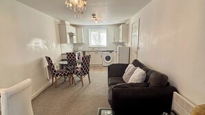A compact living space with a sofa, a glass coffee table, a dining area with patterned chairs, a washing machine, and a kitchen area. - 2 Bed Wolverhampton Apartment-sleeps 5 (Wolverhampton)