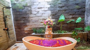 A spa area with a large oval hot tub filled with colorful flower petals, a stone wall, and a shower area. - Nidhi serene villa ubud (Kabupaten Gianyar)