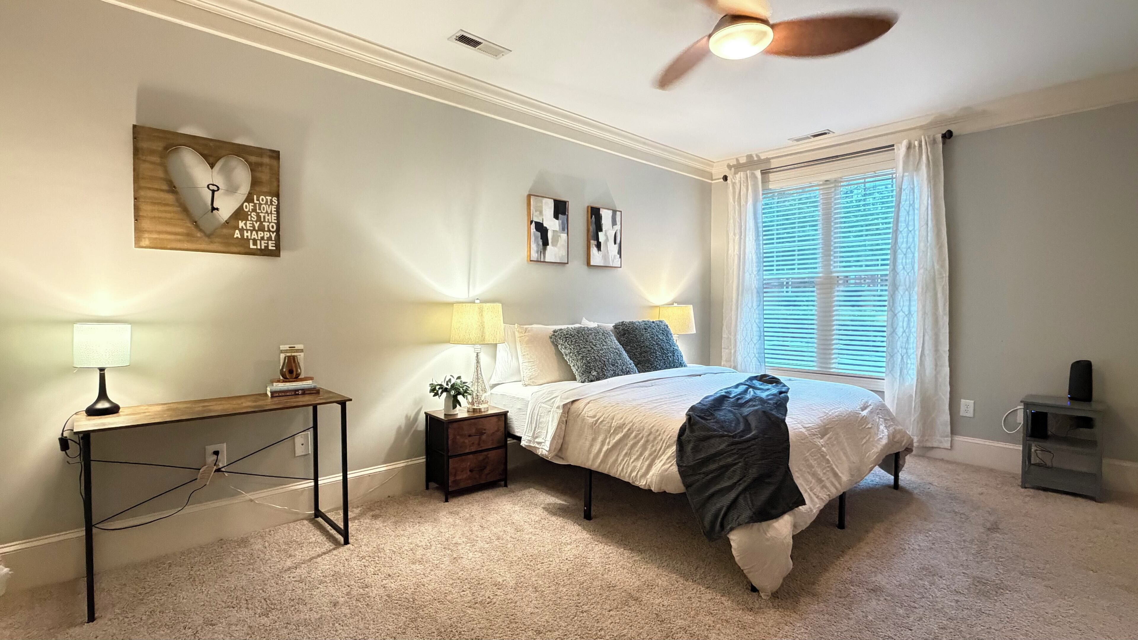 A bedroom with a bed, bedside lamps, a nightstand, a desk, a ceiling fan, and a window with curtains.