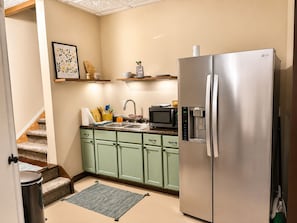 Une cuisine compacte équipée d’un réfrigérateur en acier inoxydable, d’un four à micro-ondes et d’un évier. Les meubles de rangement sont verts et un petit tapis recouvre le sol.
