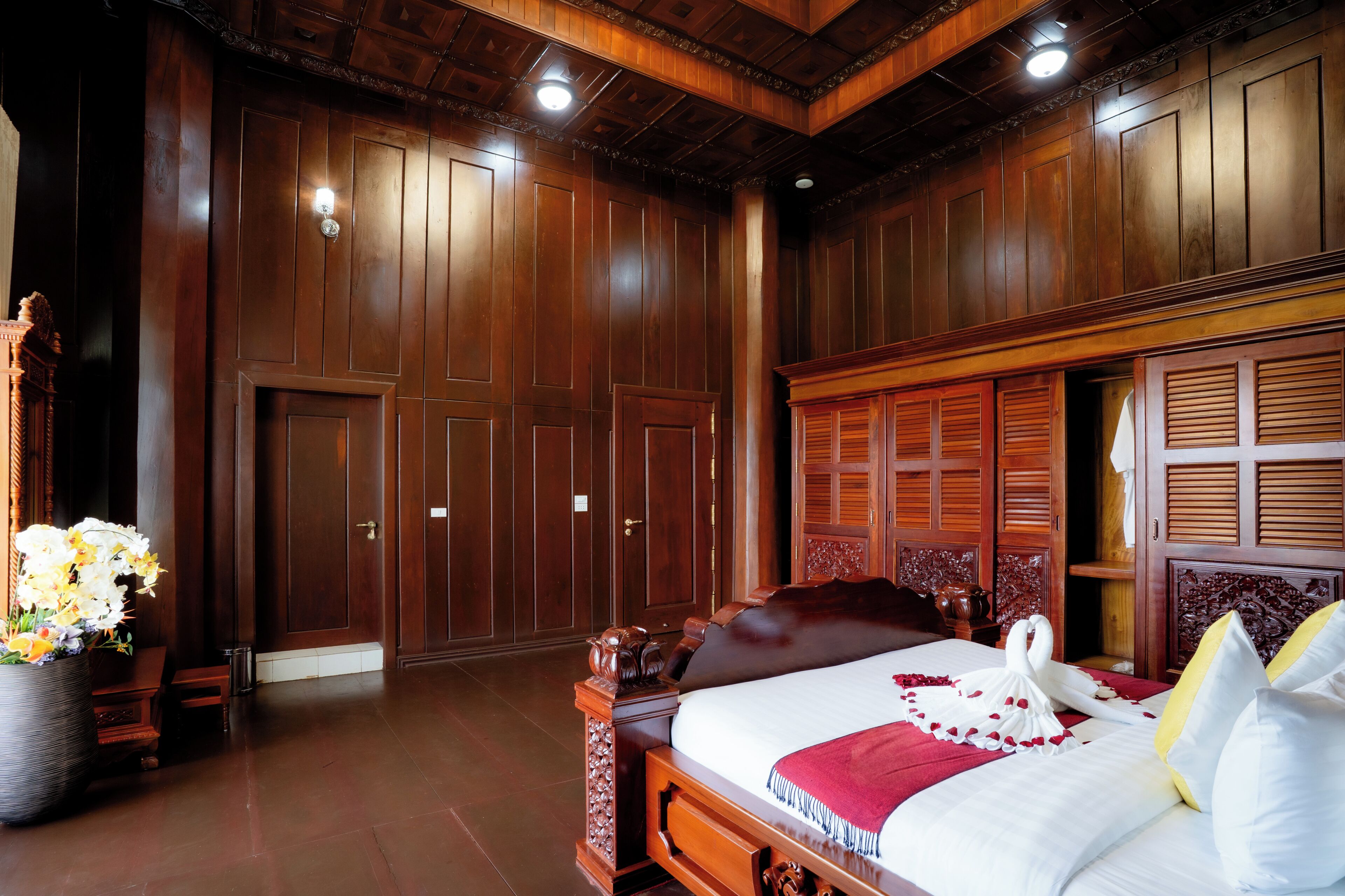 A wooden-paneled room with a large bed, a nightstand, and a vase of flowers.