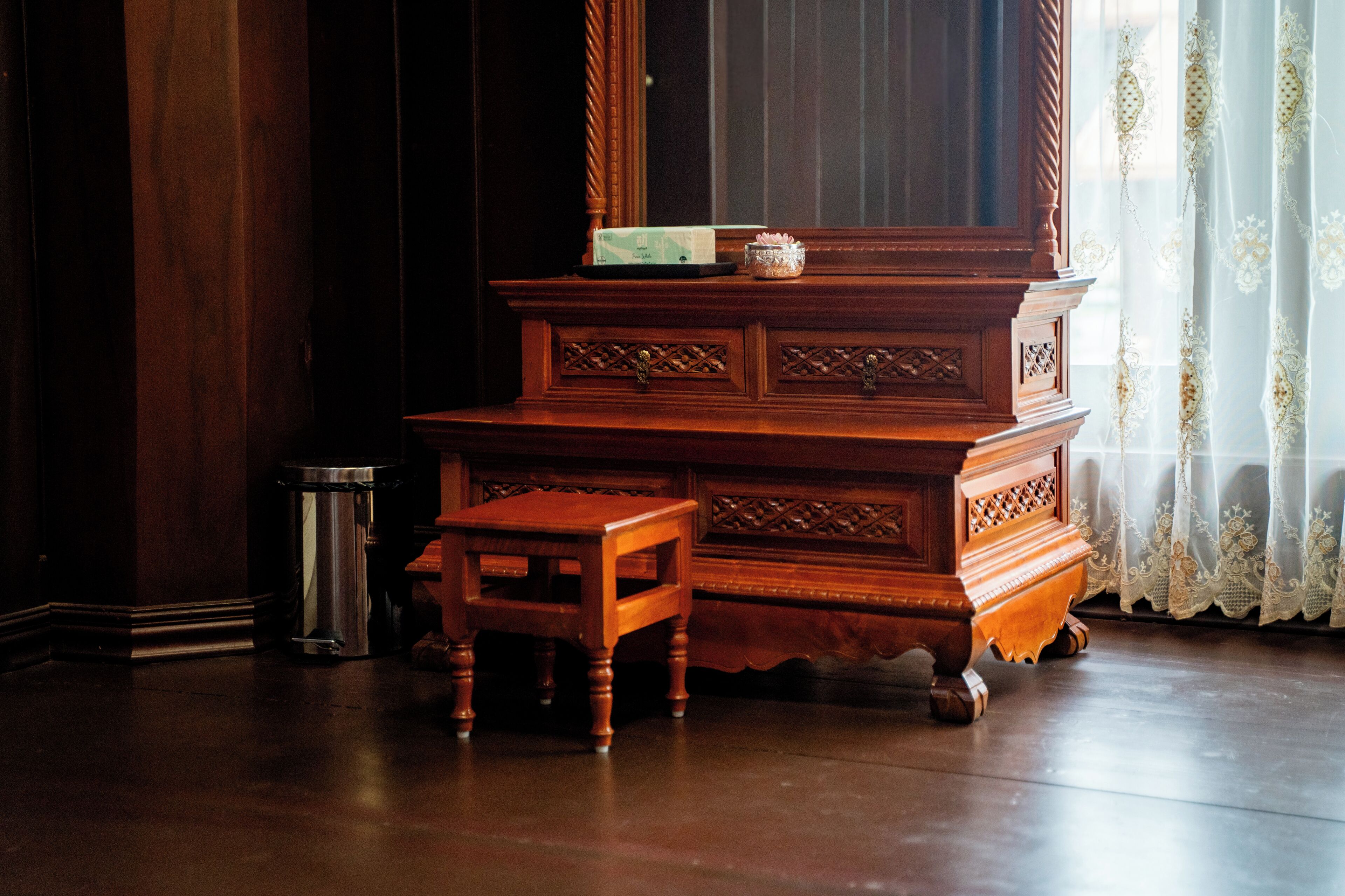 A wooden dresser with drawers, a small wooden stool, and a trash can in a room with a window and curtains.