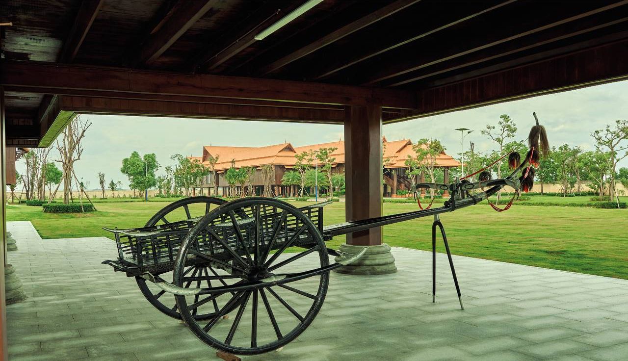 A covered outdoor area with a large wooden structure, a traditional cart, and a view of buildings in the background.
