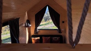 Una habitación de cabaña de madera con una cama, una ventana con cortinas, un estante con un libro y una lámpara, y vista a un paisaje cubierto de césped.