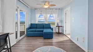 Une pièce avec un canapé bleu, un ventilateur de plafond, une vue sur l’océan par une fenêtre et un plancher en bois.