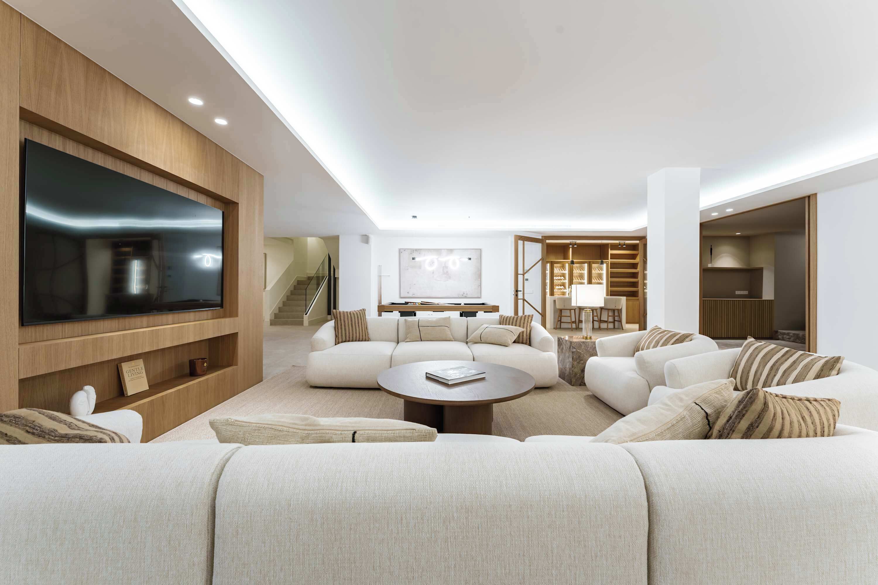 A modern living room with a large flat-screen TV, a sofa set, a coffee table, and a staircase leading to an upper level.