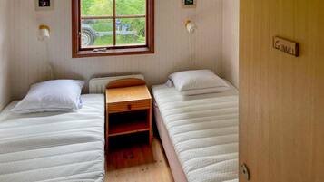 A small room with two single beds, a wooden nightstand, a window with a view of greenery, and a door labeled 'GUNN'.
