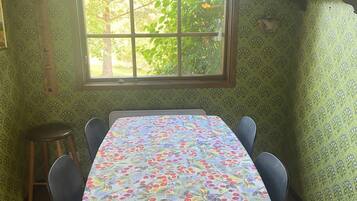 A room with a table covered in a floral tablecloth, a window with a view of greenery, and patterned wallpaper.