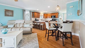 A modern kitchen with wooden cabinets, a dining area with a white table and chairs, and a living room with a beige sofa and a white coffee table. - Rooftop Tide | Ocean Views | Gas Fireplace (Salisbury)