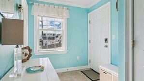 A bathroom with a white vanity, a window with a view of buildings, a white door, and a white bench. - Rooftop Tide | Ocean Views | Gas Fireplace (Salisbury)
