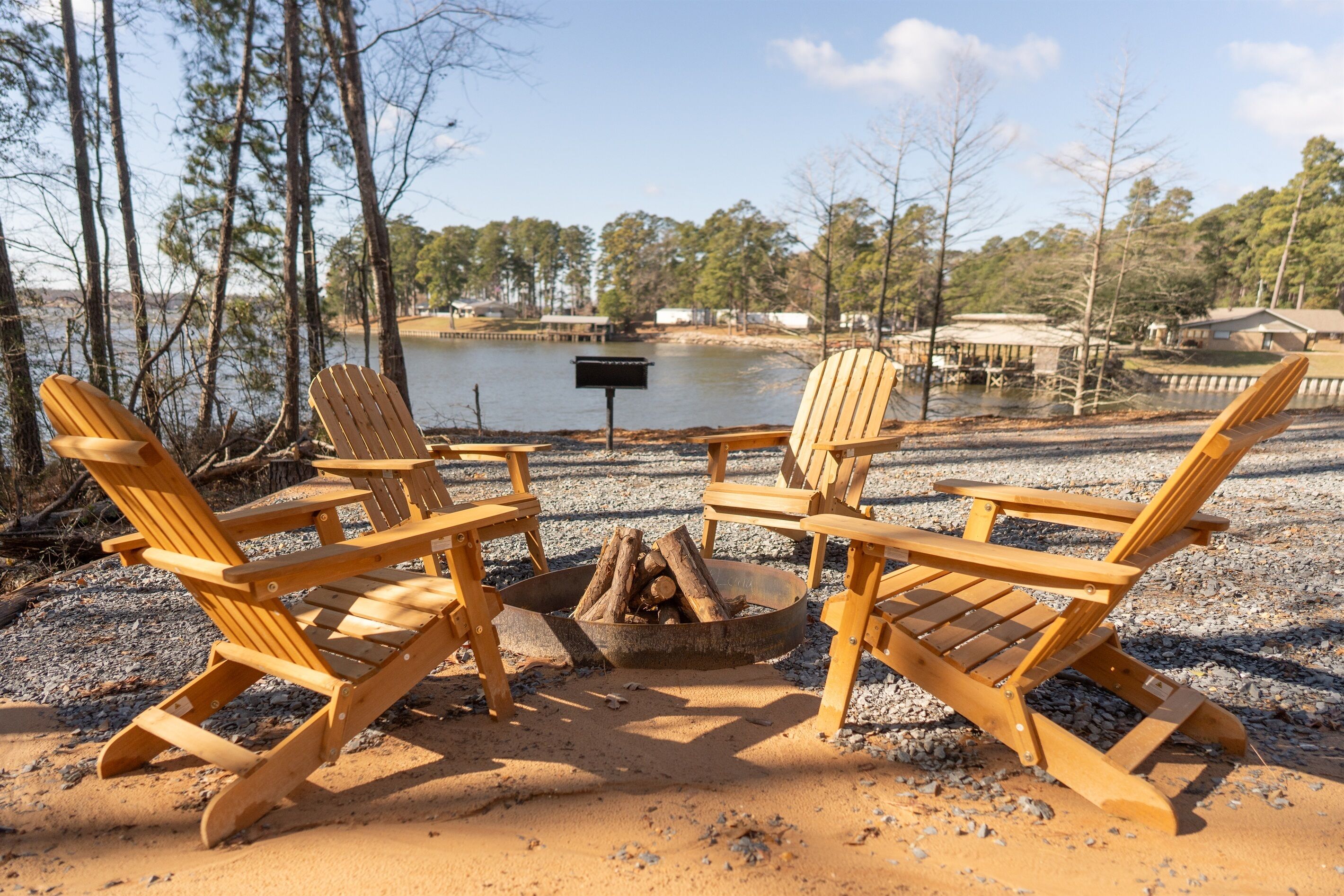 Two wooden Adirondack chairs facing a fire pit with logs, set on a gravel area with a lake and trees in the background.