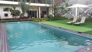 A rectangular swimming pool with a tiled edge, surrounded by a small lawn and two lounge chairs with umbrellas. - Hotel Wisma Syariah (Jakarta)