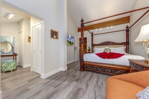 A spacious bedroom with a four-poster bed, a vanity with a mirror, and a large window.