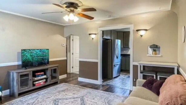 A living room with a television, a ceiling fan, a fireplace, and a kitchen in the background. - Sweet & Petite Midtown Cottage for 5-Long Stays Welcome (Billings)