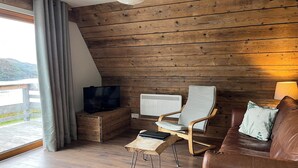 Una habitación acogedora con techo inclinado de madera, un sofá de cuero marrón, una mesa de centro de madera, un radiador blanco y un televisor sobre un mueble de madera.