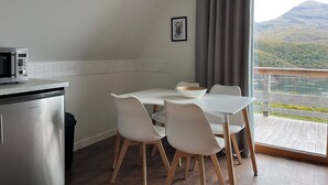 Cocina compacta con microondas, un refrigerador pequeño y un comedor con una mesa y sillas blancas. Un ventanal grande ofrece vistas de un paisaje montañoso.