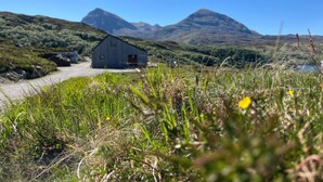 A rustic wooden cabin with a simple, functional design, surrounded by lush green hills and mountains in the background.