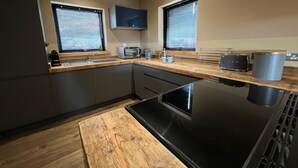 A modern kitchen with a built-in oven, microwave, and toaster. There are two windows with blinds, a sink, and a wooden countertop.