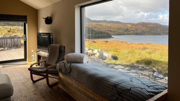 A bedroom with a large window overlooking a lake and hills, a bed, a chair, and a TV.
