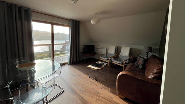 A modern living room with a glass dining table, two armchairs, a sofa, and a TV. A large window offers a view of a body of water and hills. - 3 bedroom accommodation in Kylesku, near Lairg (Kylesku, near Lairg)