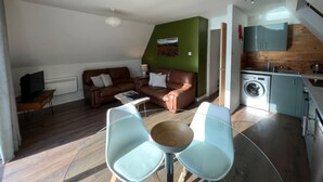 Un espacio habitable compacto con un sofá de cuero marrón, una mesa de centro de vidrio y una zona de comedor con dos sillas azul claro. Se ve una lavadora en la esquina, y una pared de acento verde aporta color a la habitación.