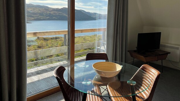 A room with a glass table and chairs, a wooden chair, a television on a stand, and a view of a lake and mountains through large windows. - Steall Gorge - UK50495 (Kylesku, near Lairg)