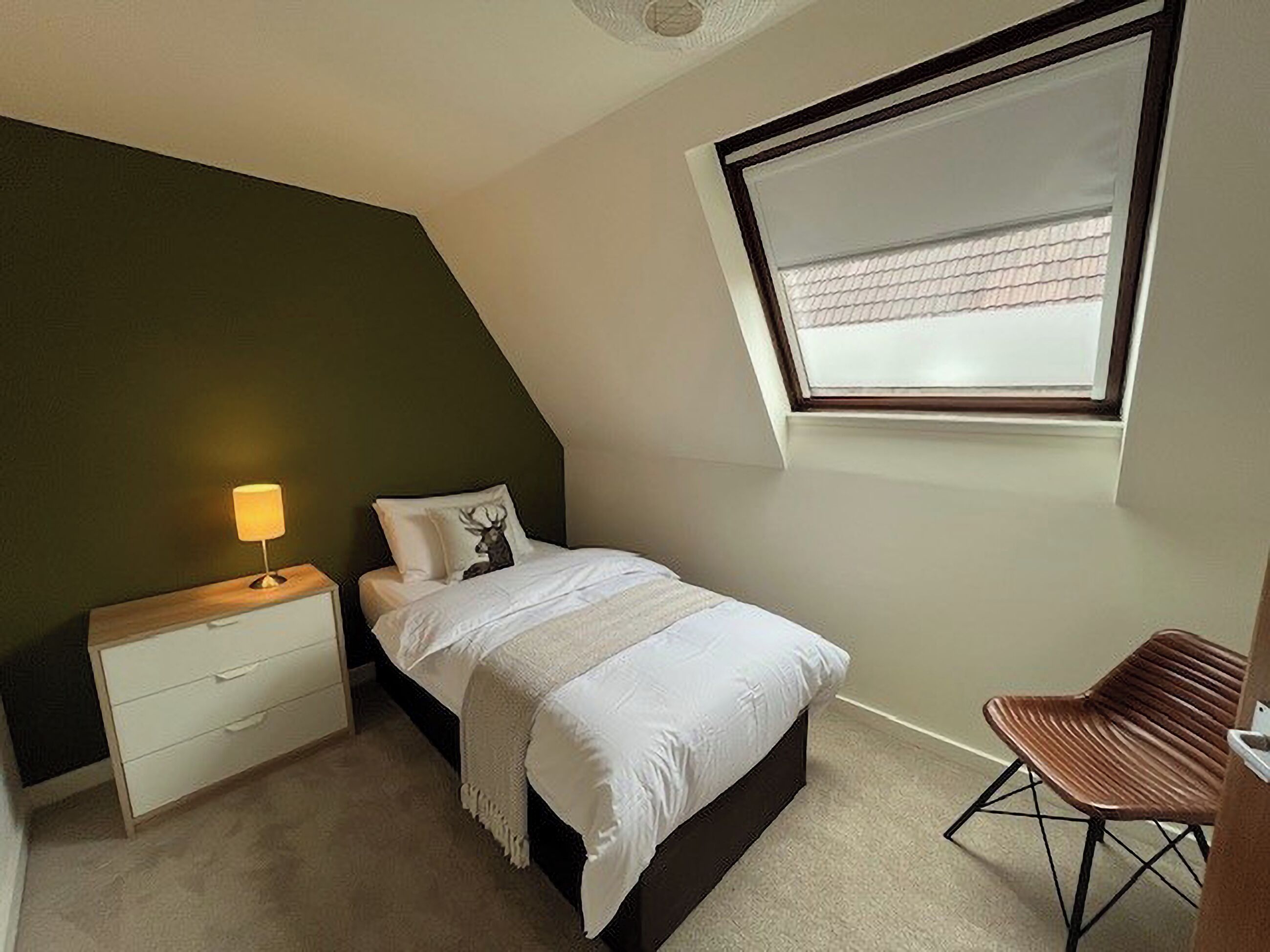 Un dormitorio con techo inclinado, una cama con cubrecama blanco, una mesita de noche con lámpara, una silla y una ventana con persianas.