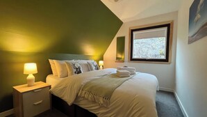 A neatly made bed with white linens and decorative pillows, a bedside table with a lamp, and a window with a view of trees. - 3 bedroom accommodation in Kylesku, near Lairg (Kylesku, near Lairg)