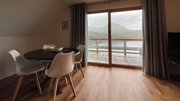 A room with a dining table, chairs, a TV, and a large window with a view of a lake and mountains. - 3 bedroom accommodation in Kylesku, near Lairg (Kylesku, near Lairg)