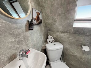 Une salle de bain avec un lavabo blanc, des toilettes et un miroir. Un tableau encadré représentant un chien est accroché au mur, et un flacon de savon liquide se trouve sur le lavabo.