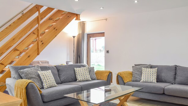 Uma sala de estar moderna com uma mesa de centro de vidro, dois sofás cinzas com almofadas estampadas, uma escada de madeira e vista através de uma porta de vidro.