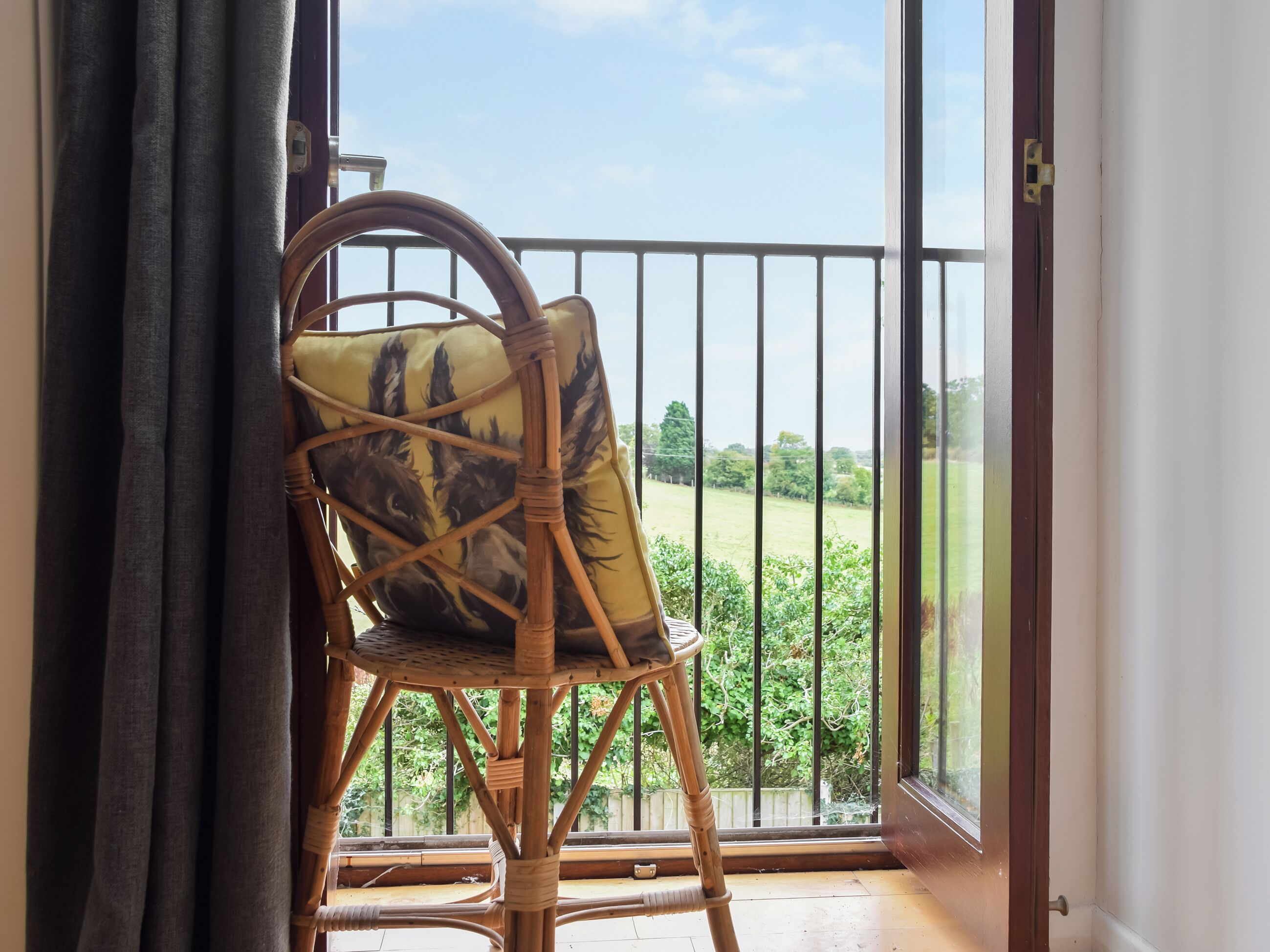Une chaise en osier, agrémentée d’un coussin à motifs, est placée sur un balcon doté d’une rambarde en métal, d’où l’on peut admirer un paysage verdoyant.