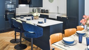 A modern kitchen with dark cabinetry, a central island with bar stools, a sink, and a clock on the wall. - 4 bedroom accommodation in Little Dunham (Little Dunham)