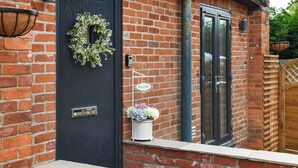 A brick building with a dark door, a welcome sign, a hanging light fixture, and a potted plant. - 4 bedroom accommodation in Little Dunham (Little Dunham)