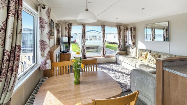 A spacious living room with a wooden dining table, chairs, a sofa, and a large window offering a view of a residential area. - 3 bedroom accommodation in California Cliffs (California Cliffs)