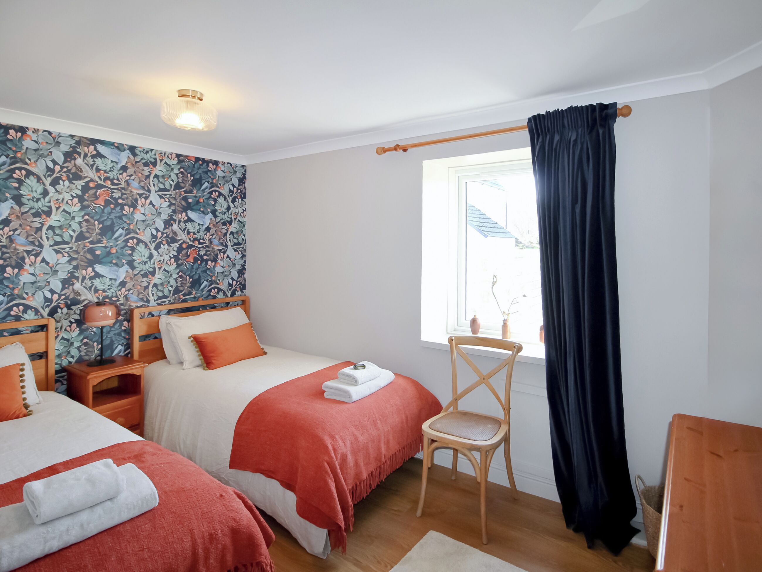 Une chambre avec deux lits, une chaise, une fenêtre avec des rideaux, une lampe et un mur orné d’un motif floral.