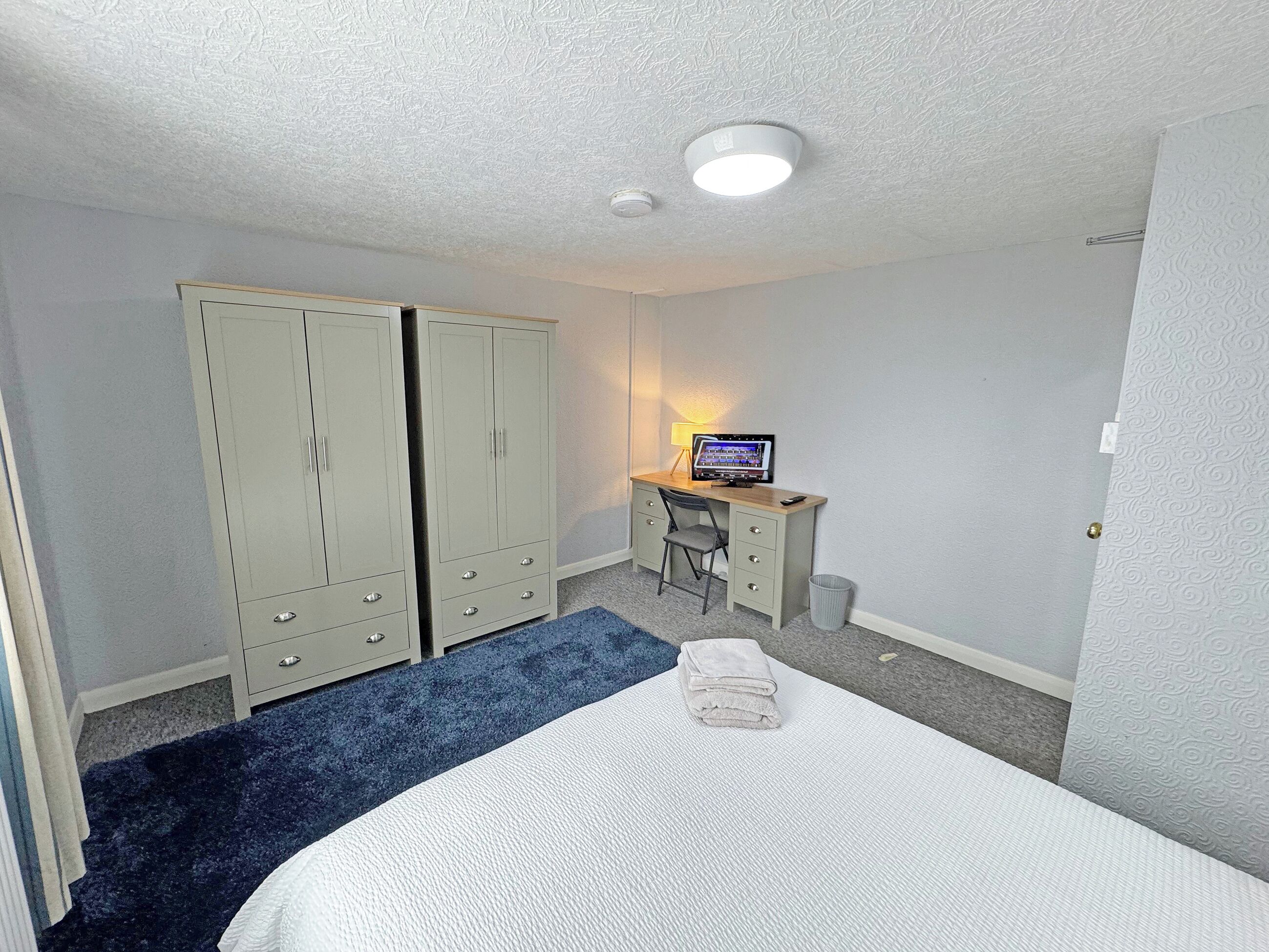 Een slaapkamer met een bed, een bureau met een computer, twee kasten en een kleine prullenbak.