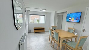 A room with a wooden table and chairs, a television on the wall, a window with a view of a brick building, and a small bench.