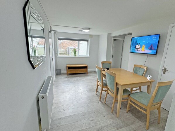 A room with a wooden table and chairs, a television on the wall, a window with a view of a brick building, and a small bench.