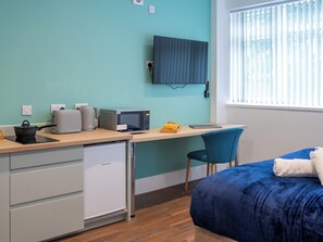 A compact room with teal walls, a desk with a microwave, a small refrigerator, a bed with a blue blanket, and a window with blinds.