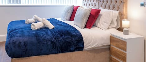 Una cama bien hecha con una colcha azul y toallas enrolladas, un cabecero capitonado y una mesita de noche con una lámpara.