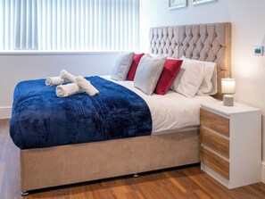 A neatly made bed with a blue blanket and rolled towels, a tufted headboard, and a bedside table with a lamp.