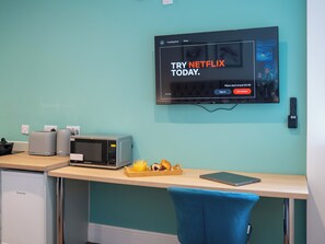 A modern kitchen with a microwave, toaster, and kettle on the counter, a small refrigerator, and a TV mounted on the wall displaying a Netflix advertisement.