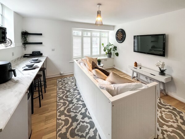 A modern kitchen with a dining area, a TV mounted on the wall, and a window with blinds.