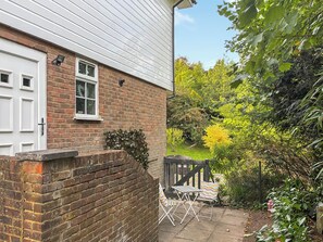 A brick house with a white door, a small window, and a garden with a wooden fence and chairs. - A lovely cosy property in a lovely location with stunning scenery. (Kenley, near Surrey)